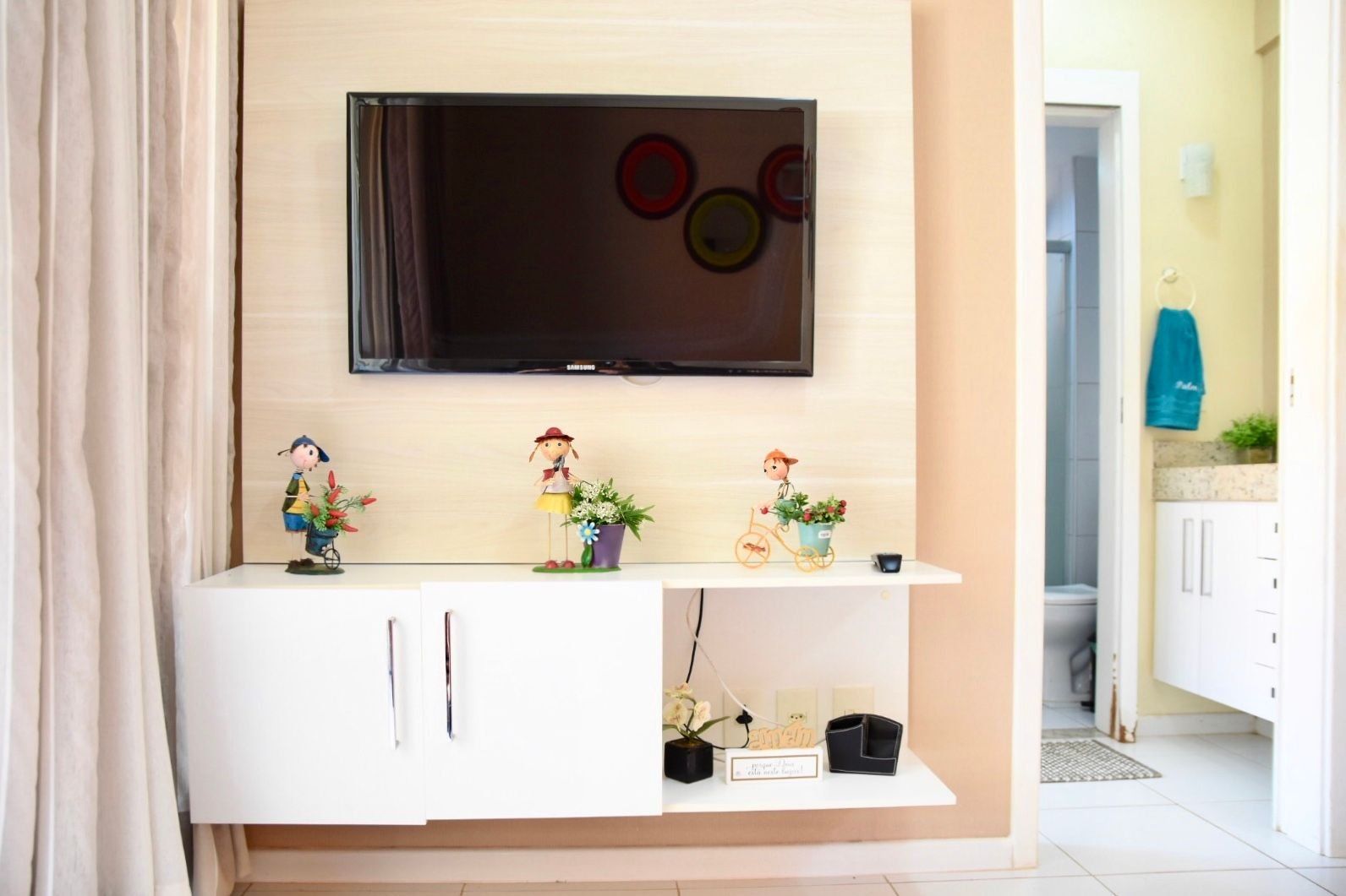 A modern living room with a flat-screen TV mounted on the wall, a white entertainment unit with decorative figurines, and a doorway leading to a bathroom.