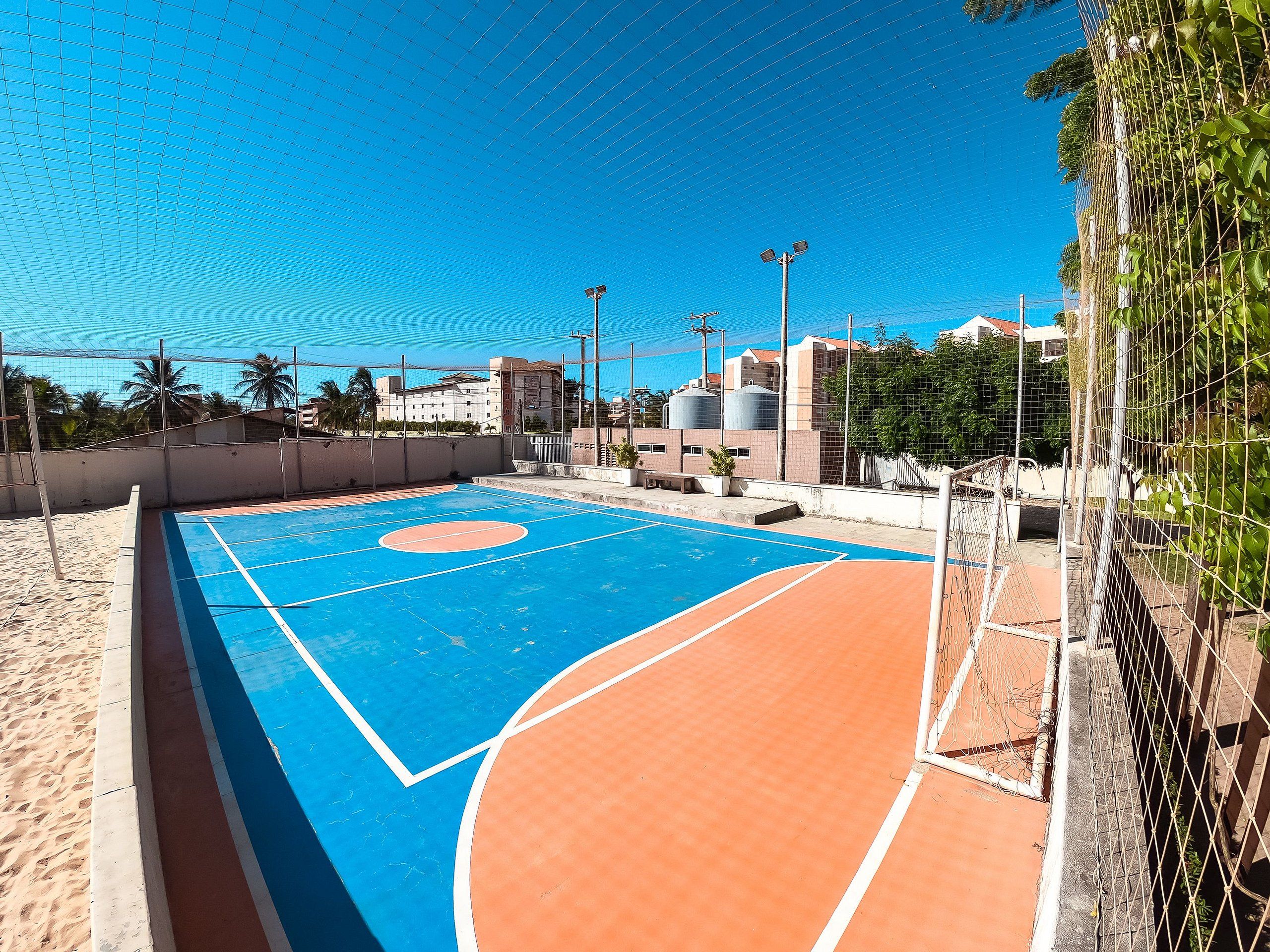 Una cancha de baloncesto al aire libre con superficie azul y naranja, rodeada por una cerca de alambrón, y edificios al fondo.