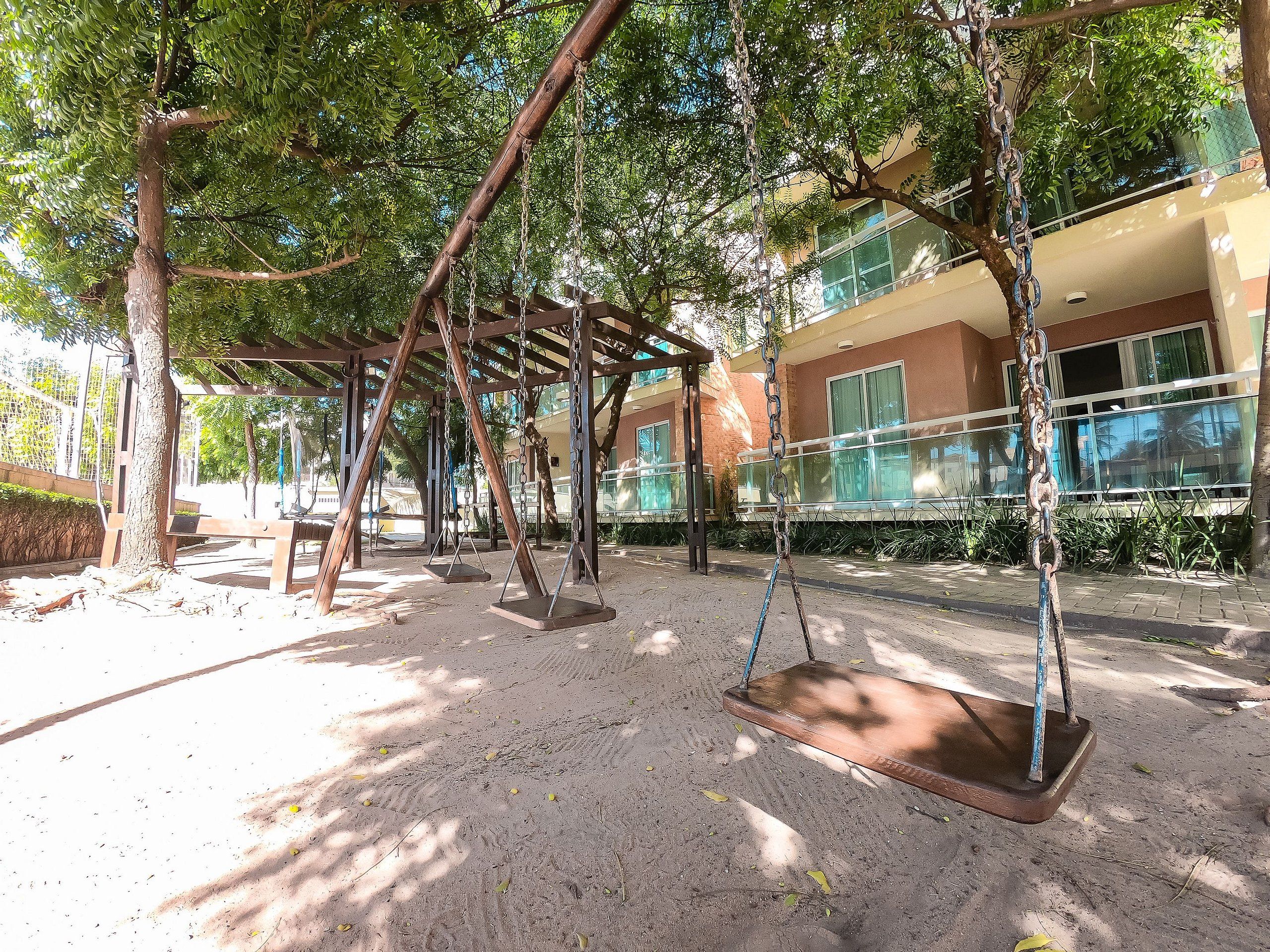 A playground with swings and a covered seating area, surrounded by trees and a building with large windows.