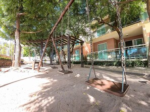 A playground with swings and a covered seating area, surrounded by trees and a building with large windows.