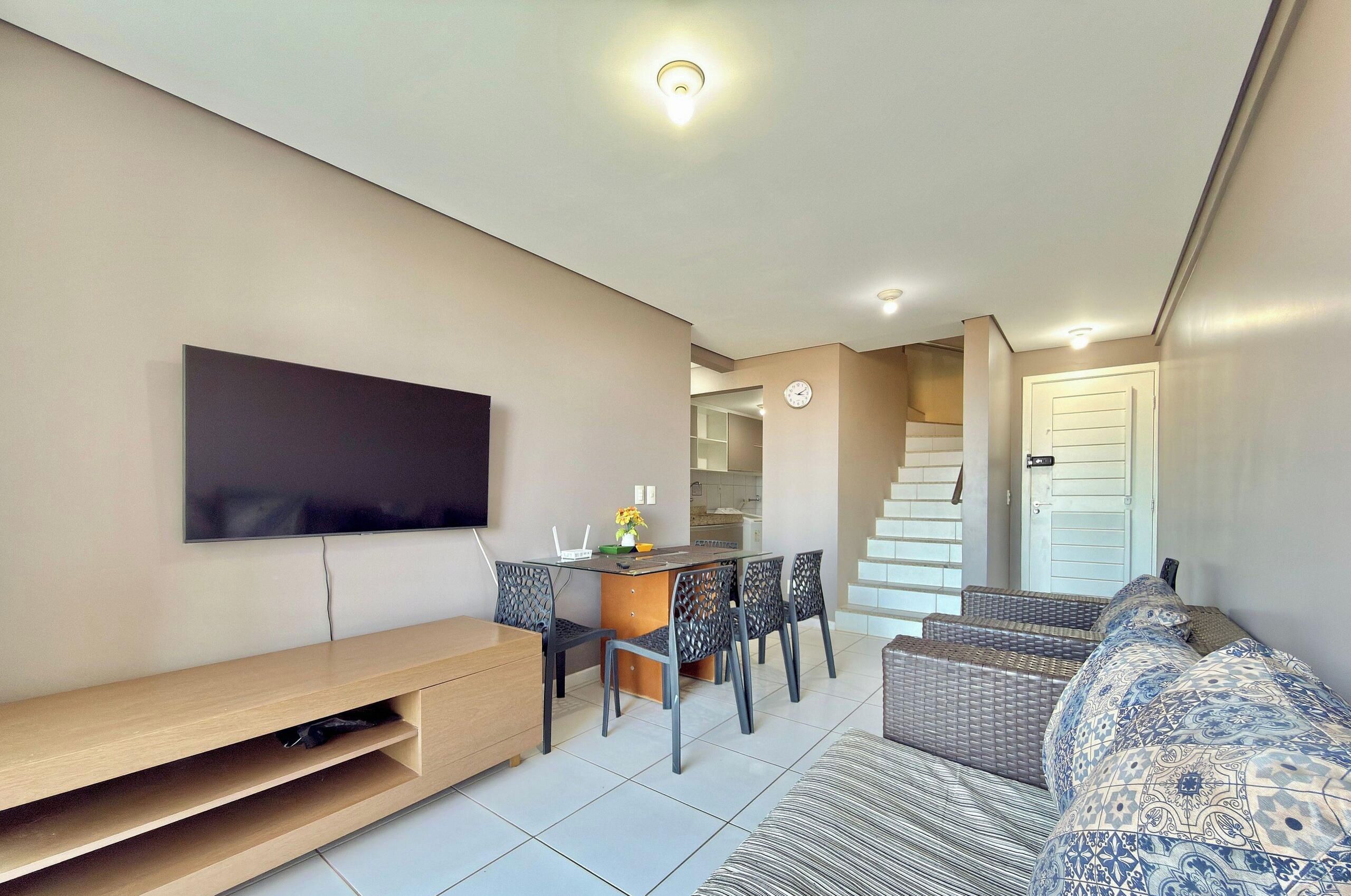 Una sala de estar moderna con un televisor de pantalla plana, un comedor con mesa y sillas, y una escalera que conduce a un nivel superior.