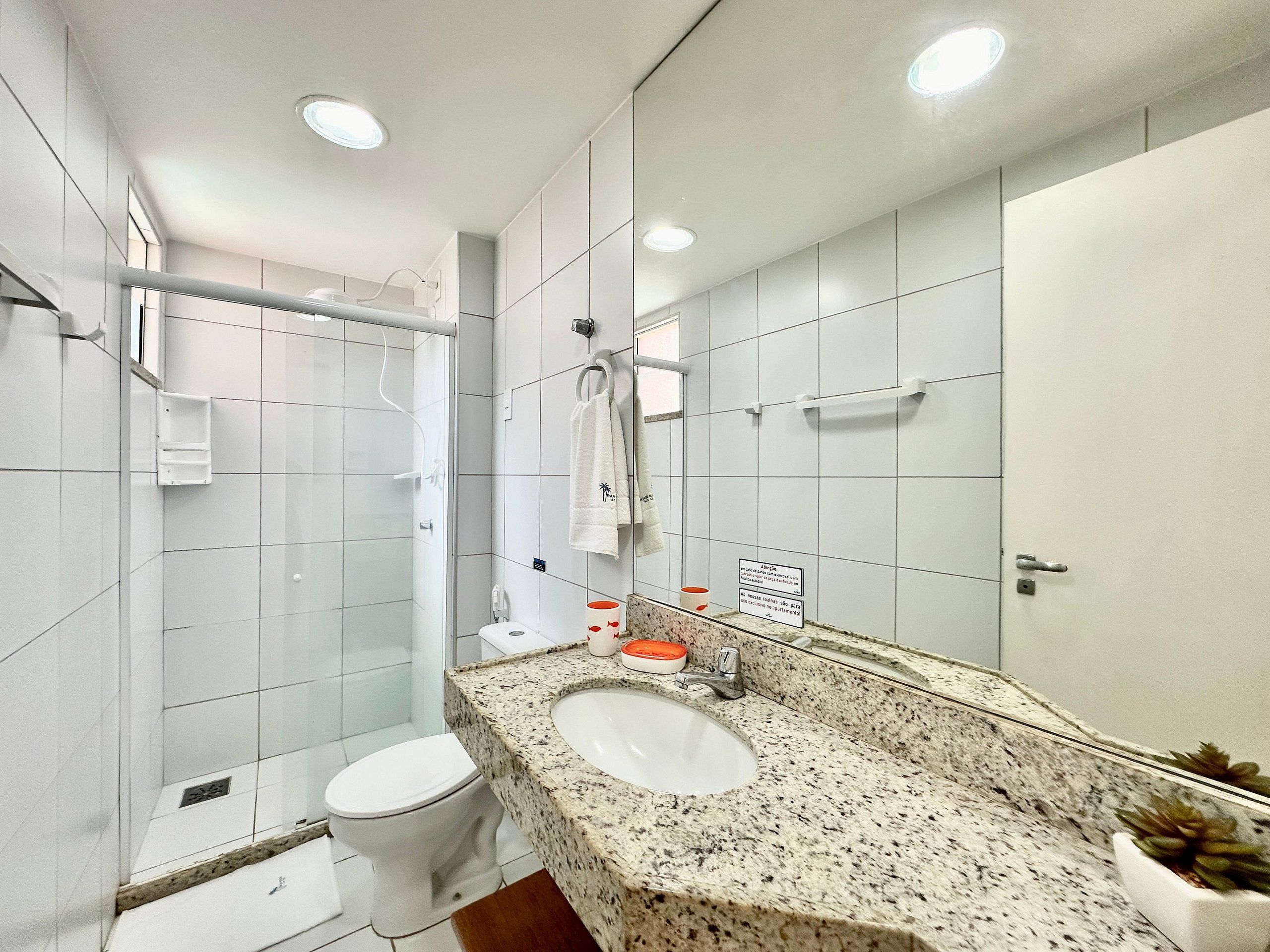 Un baño moderno con cabina de ducha de vidrio, inodoro blanco, lavamanos con encimera de granito y un espejo grande.