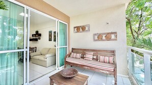 En balkon med træbænk, bord og en glasdør, der fører til en opholdsstue med sofa og spisebord.