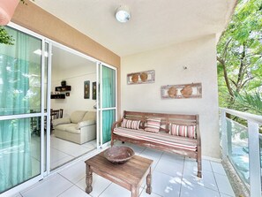 A balcony with a wooden bench, a table, and a glass door leading to a living area with a sofa and a dining table. - Palm Beach Porto das Dunas - CE (Aquiraz)