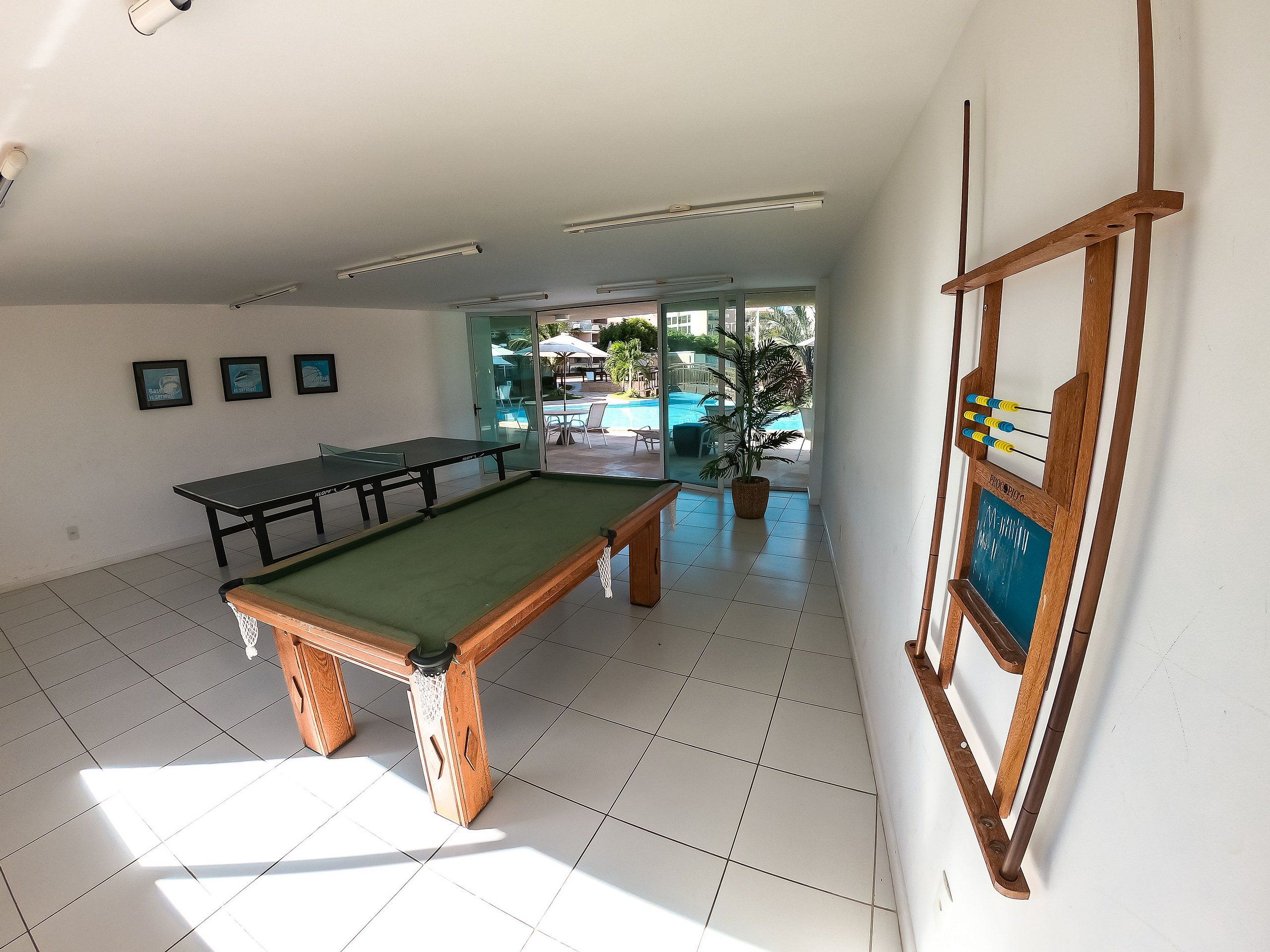 Mesa de bilhar numa sala com chão de azulejos, teto com luzes fluorescentes e vista para uma piscina ao ar livre através de uma porta de vidro.