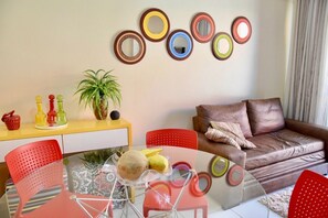 Een woonkamer met een glazen tafel, rode stoelen, een bruine leren bank, een geel kastje en een muur met ronde, kleurrijke spiegels.