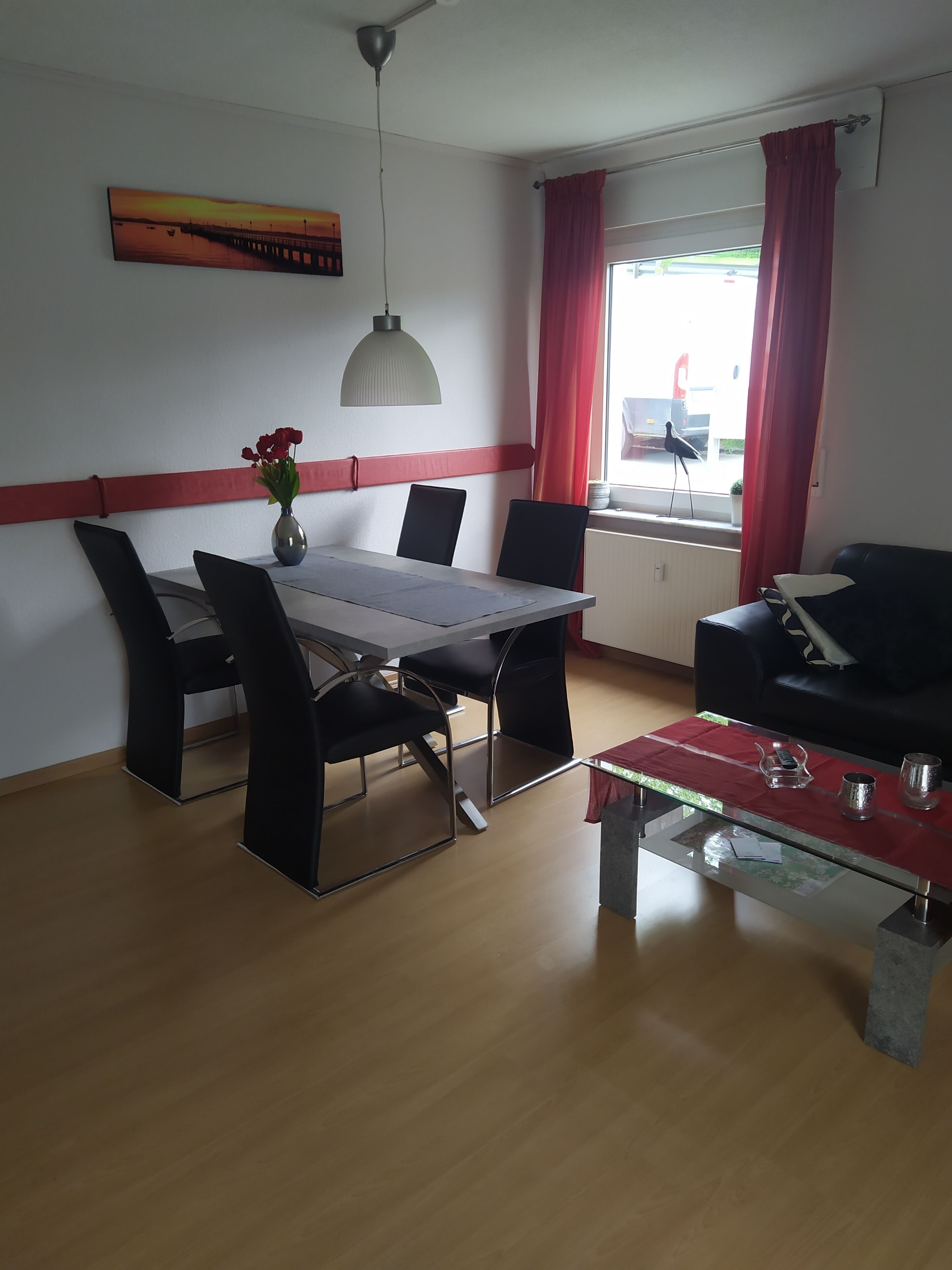 Ein Essbereich mit Tisch, Stühlen, einer Blumenvase, einer Pendelleuchte und Blick ins Freie durch ein Fenster mit roten Vorhängen.