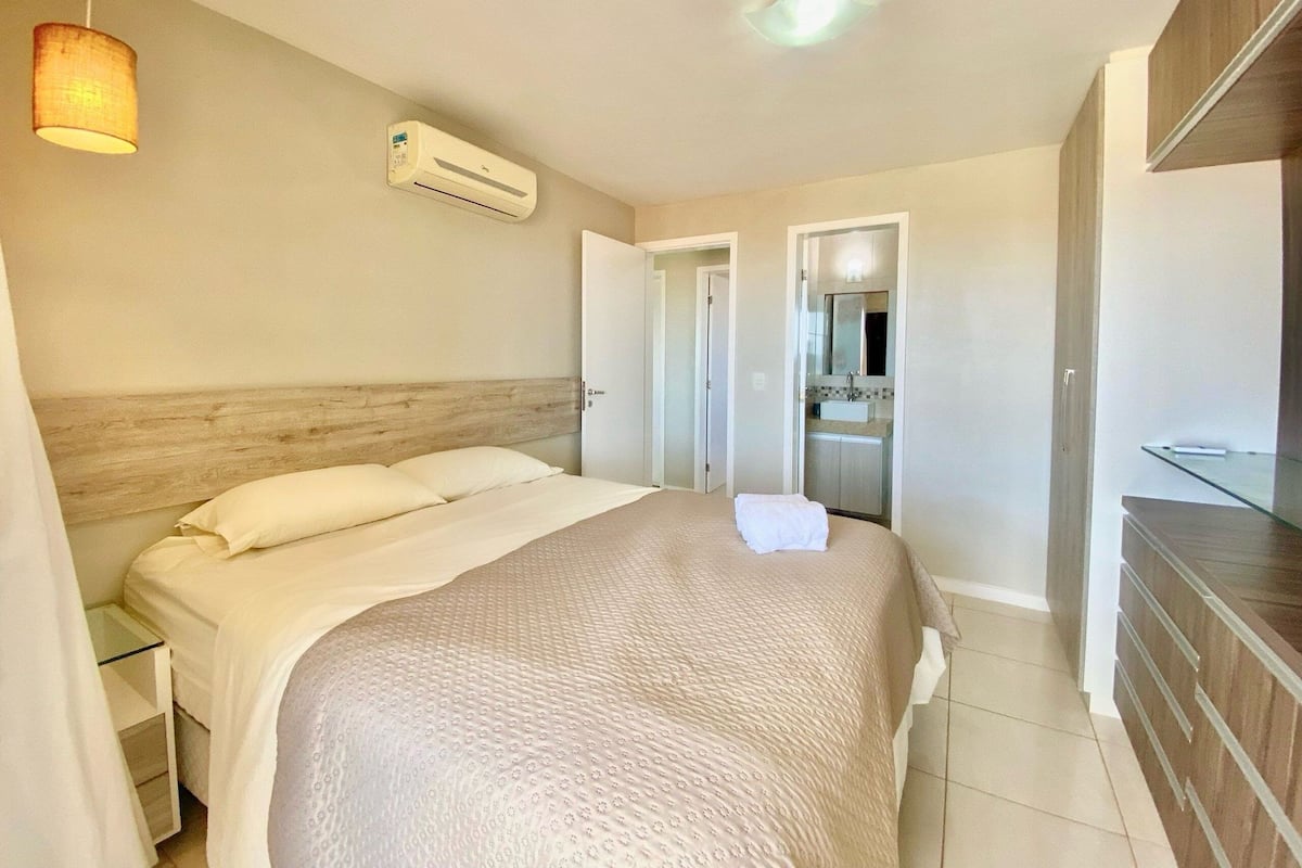 A bedroom with a bed, an air conditioner, a bedside table, and a mirror in the adjacent bathroom.