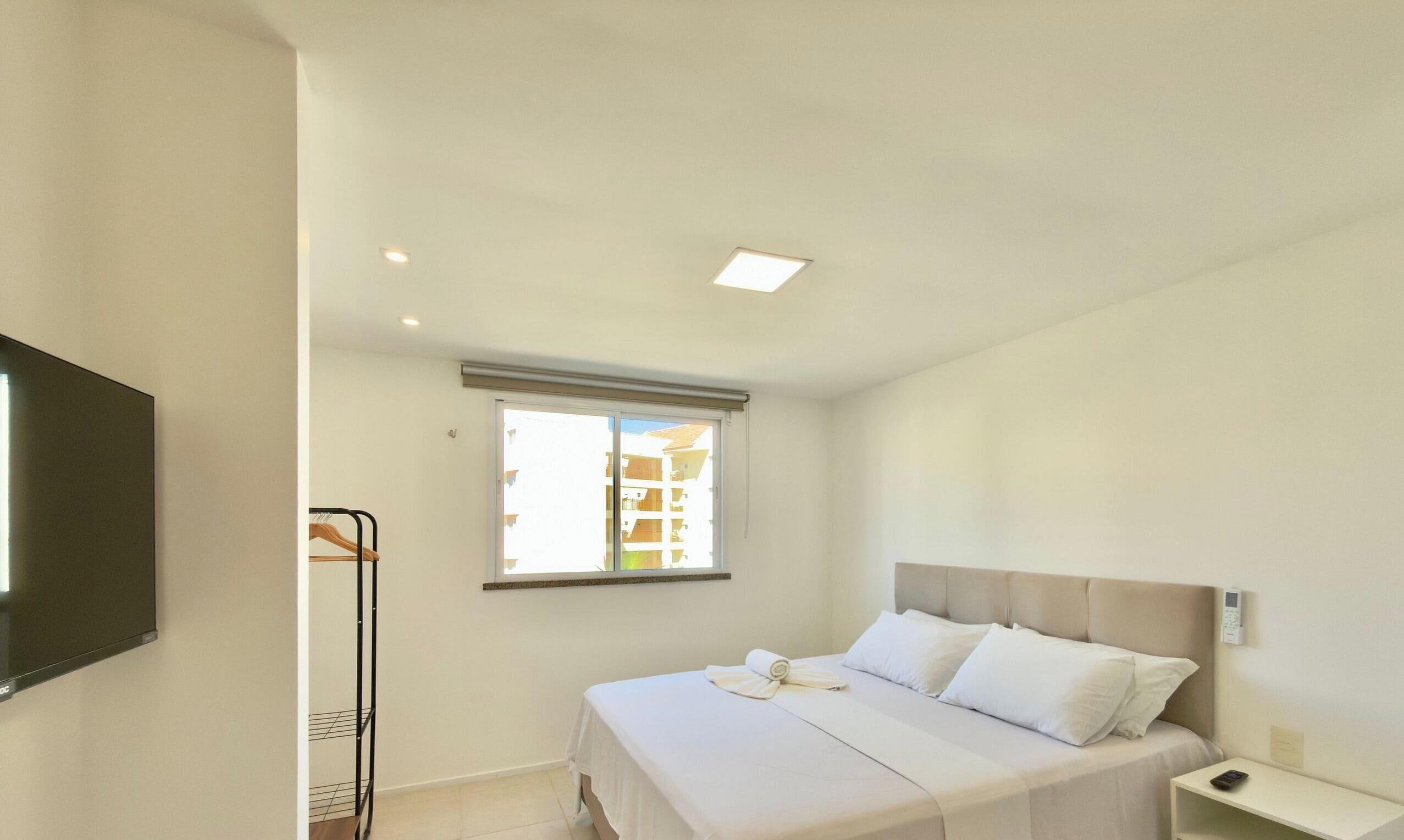 A bedroom with a bed, a bedside table, a nightstand, a window with blinds, a ceiling light, and a wall-mounted TV.