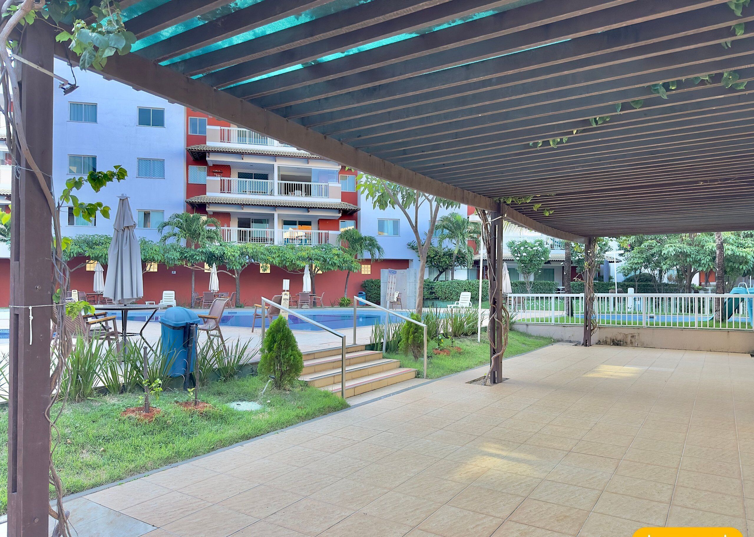 A covered outdoor area with a pool, lounge chairs, and a building with balconies in the background.