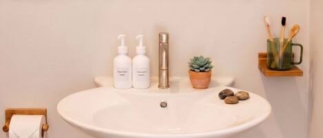 A bathroom with a white sink, two bottles of shampoo, a potted plant, and a trash bin.