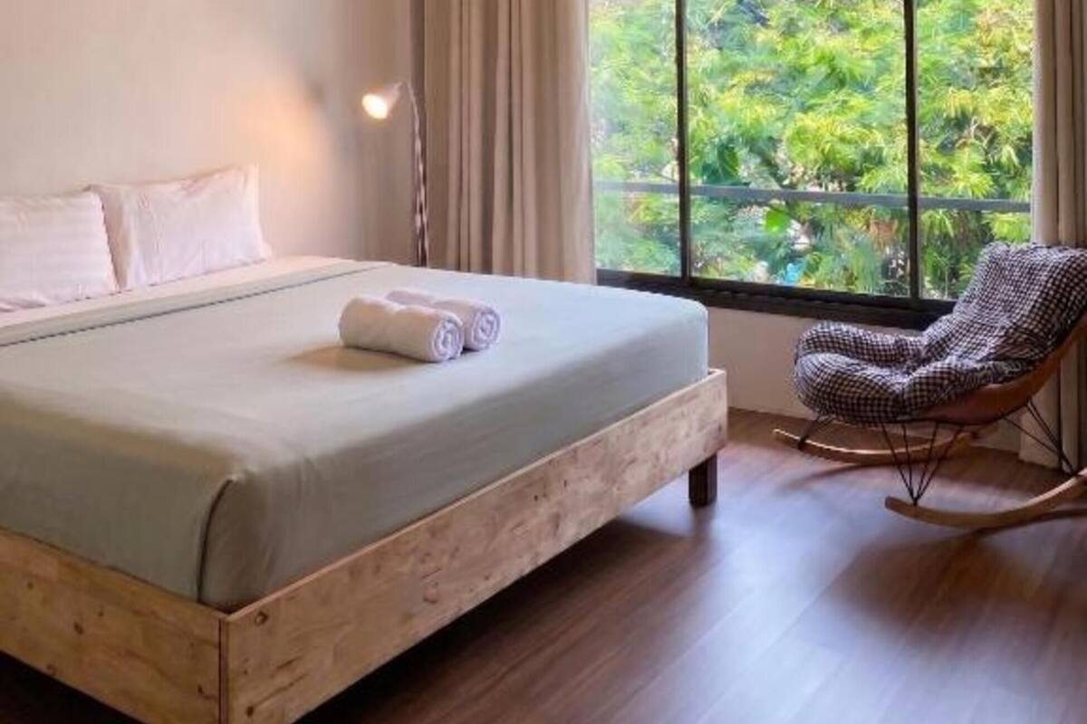 Une chambre à coucher avec un grand lit, un fauteuil à bascule, une fenêtre donnant sur un paysage verdoyant et un luminaire au plafond.