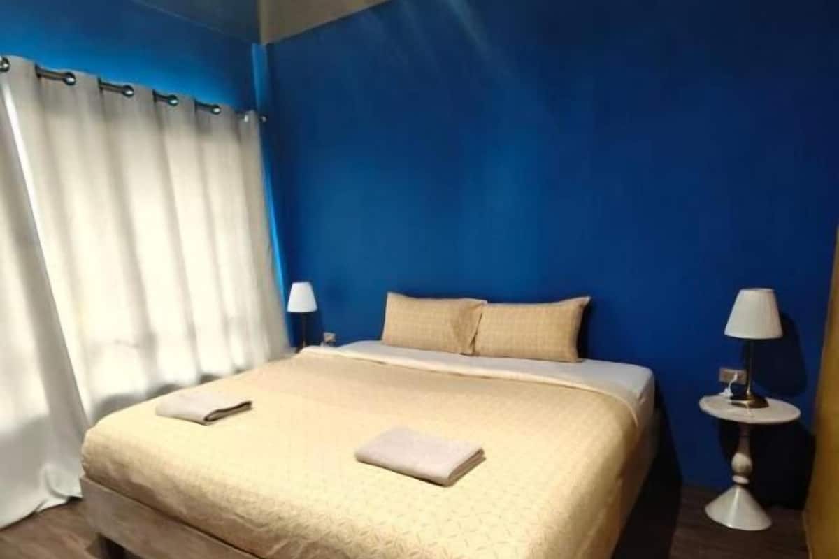 Une chambre d’hôtel avec un lit, deux tables de chevet, une petite table, un tapis rond et une fenêtre avec des rideaux.