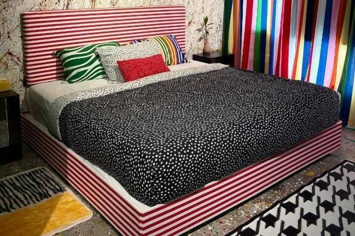 Une chambre avec un lit à rayures, une couvre-lit à motifs, un coussin à rayures zébrées, un coussin rouge, un coussin vert, un mur avec un motif texturé, une œuvre d’art encadrée, une table de chevet avec une lampe et un rideau à rayures.