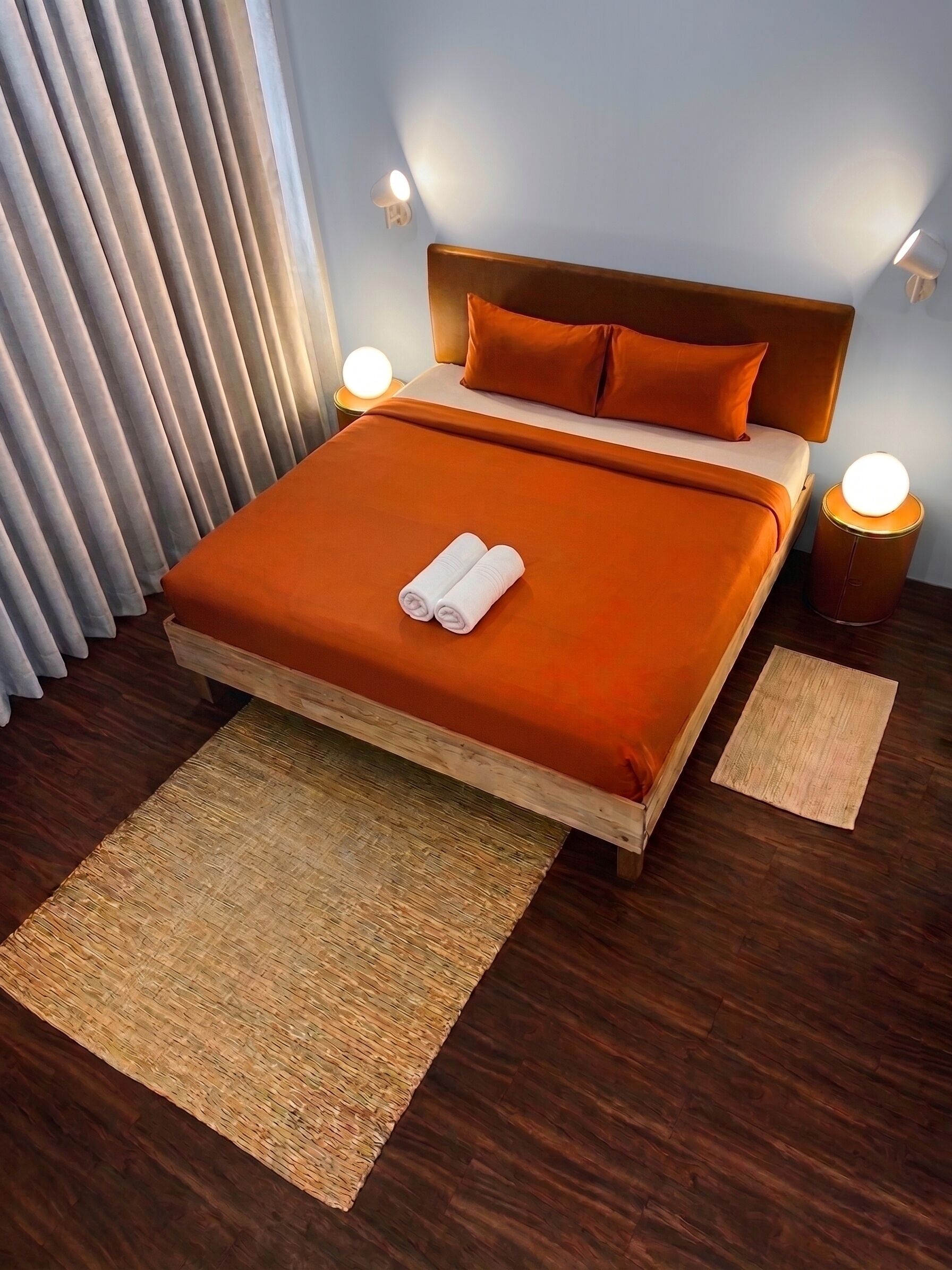 Una cama con una colcha naranja, dos almohadas y dos toallas enrolladas. Hay dos luces fijadas a la pared y una mesa redonda con una lámpara.
