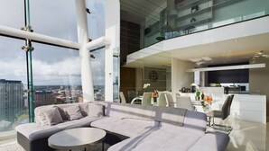 A modern living room with a large L-shaped sectional sofa, a round coffee table, and a glass dining table with chairs. The room features a large window with a city view, a kitchen area with modern appliances, and a glass railing overlooking the city. - Skyline 3BR Duplex | Pool, Gym & Parking (Manchester)