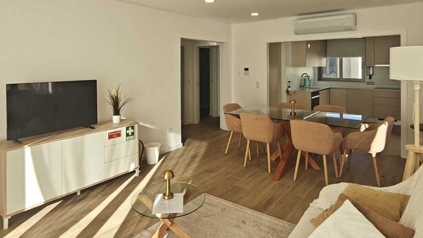 A modern living room with a flat-screen TV, a sofa, a dining table with chairs, and a kitchen area in the background. - 2 bedroom apartment 1 minute walk from Cortegaça beach - Refugio do Surf Cortegaça (Cortegaça)