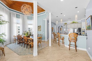 A spacious dining area with wooden chairs and a large chandelier, a kitchen with modern appliances, and a bar area with stools. - Southern Dream: Two Units (Fort Myers Beach)