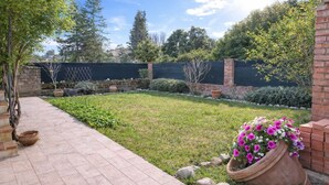 Un giardino sul retro con un vialetto lastricato, un muro in mattoni, un vaso da fiori grande e diverse piante e alberi.