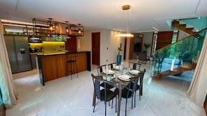 A modern kitchen with a central island, dining area with a glass table, and a staircase leading to an upper level. - Casa c Jacuzzi pr x Praia do Forte Lagoa do Aru (Mata de São João)