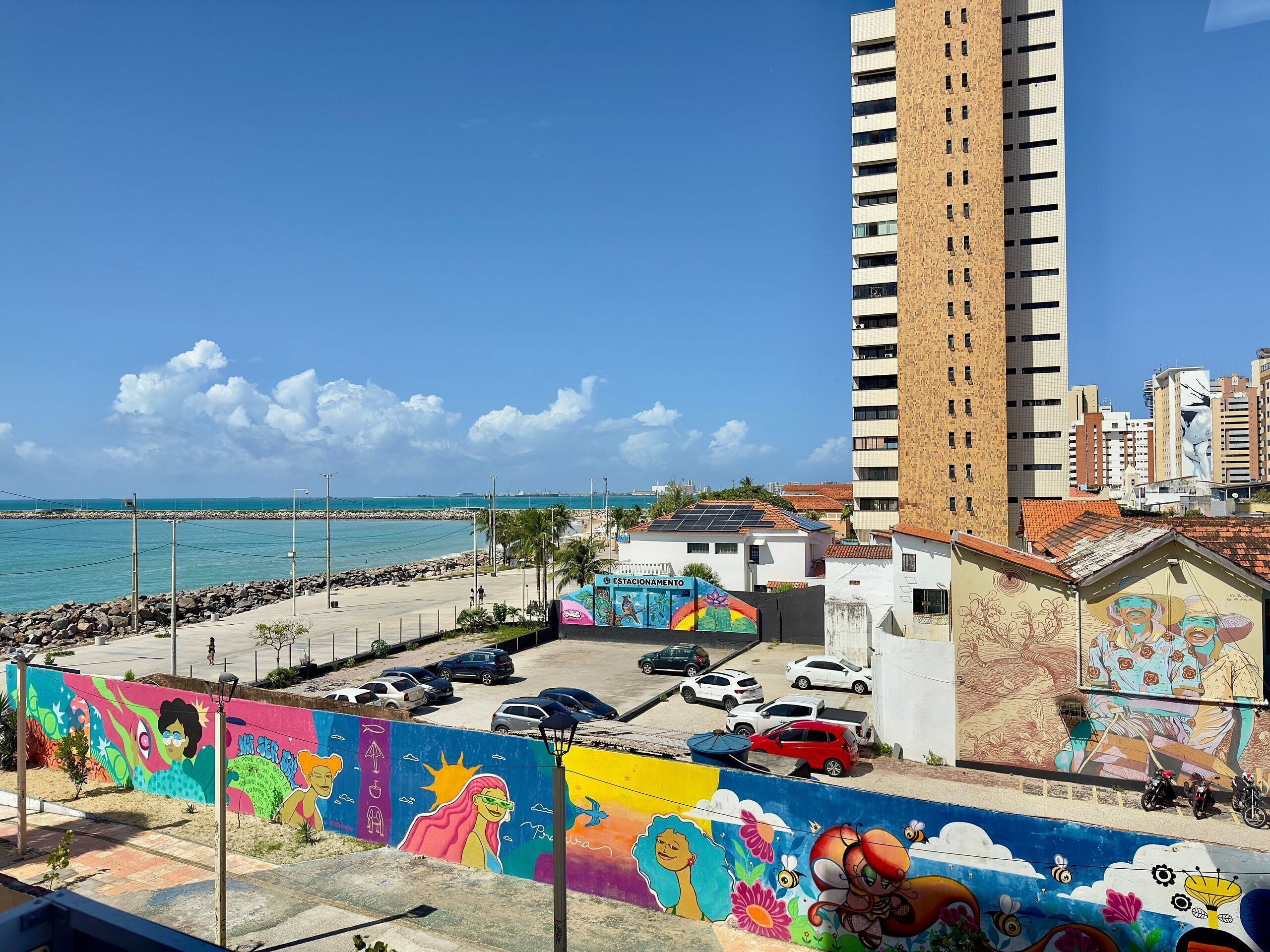A coastal cityscape with a colorful mural, a tall building, and a clear blue sky.