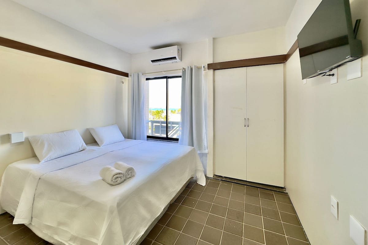 A hotel room with a bed, a TV, a sliding door, and a view of the beach.