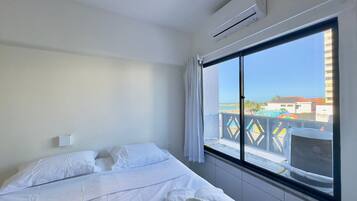 Quarto de hotel com cama, roupa de cama branca, unidade de ar condicionado, janela com vista para edifícios e uma piscina.