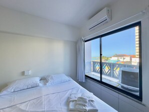 A hotel room with a bed, white linens, an air conditioning unit, a window with a view of buildings and a pool. - Flats Tabajara Residence (Fortaleza)