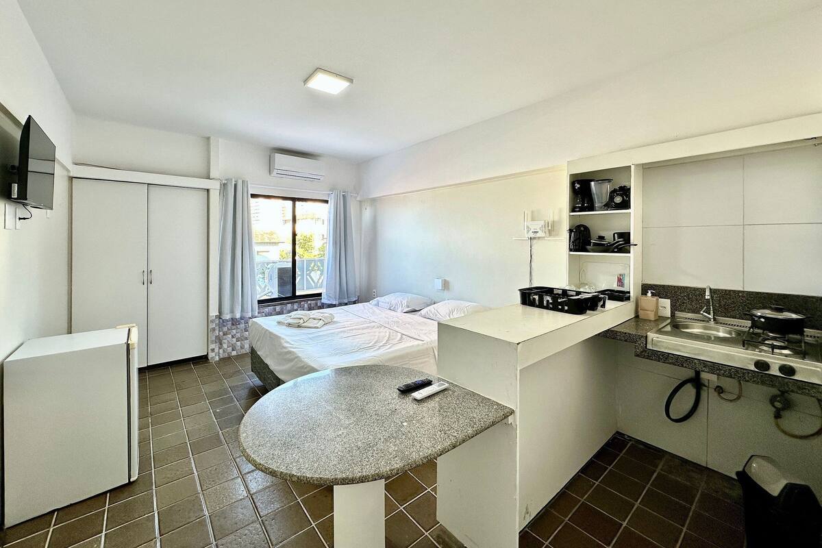 A compact hotel room with a bed, a small kitchenette, a bathroom, and a balcony with curtains.