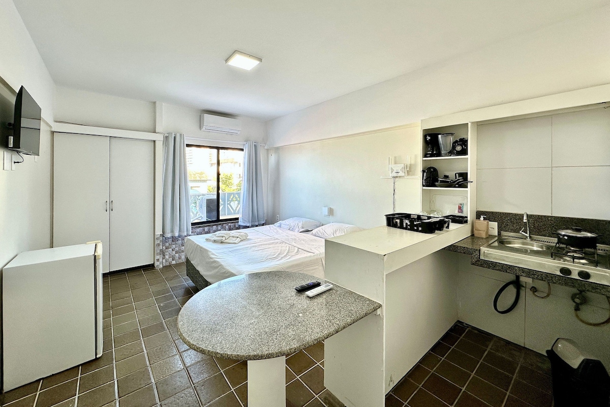 A compact hotel room with a bed, a small kitchenette, a bathroom, and a balcony with curtains.