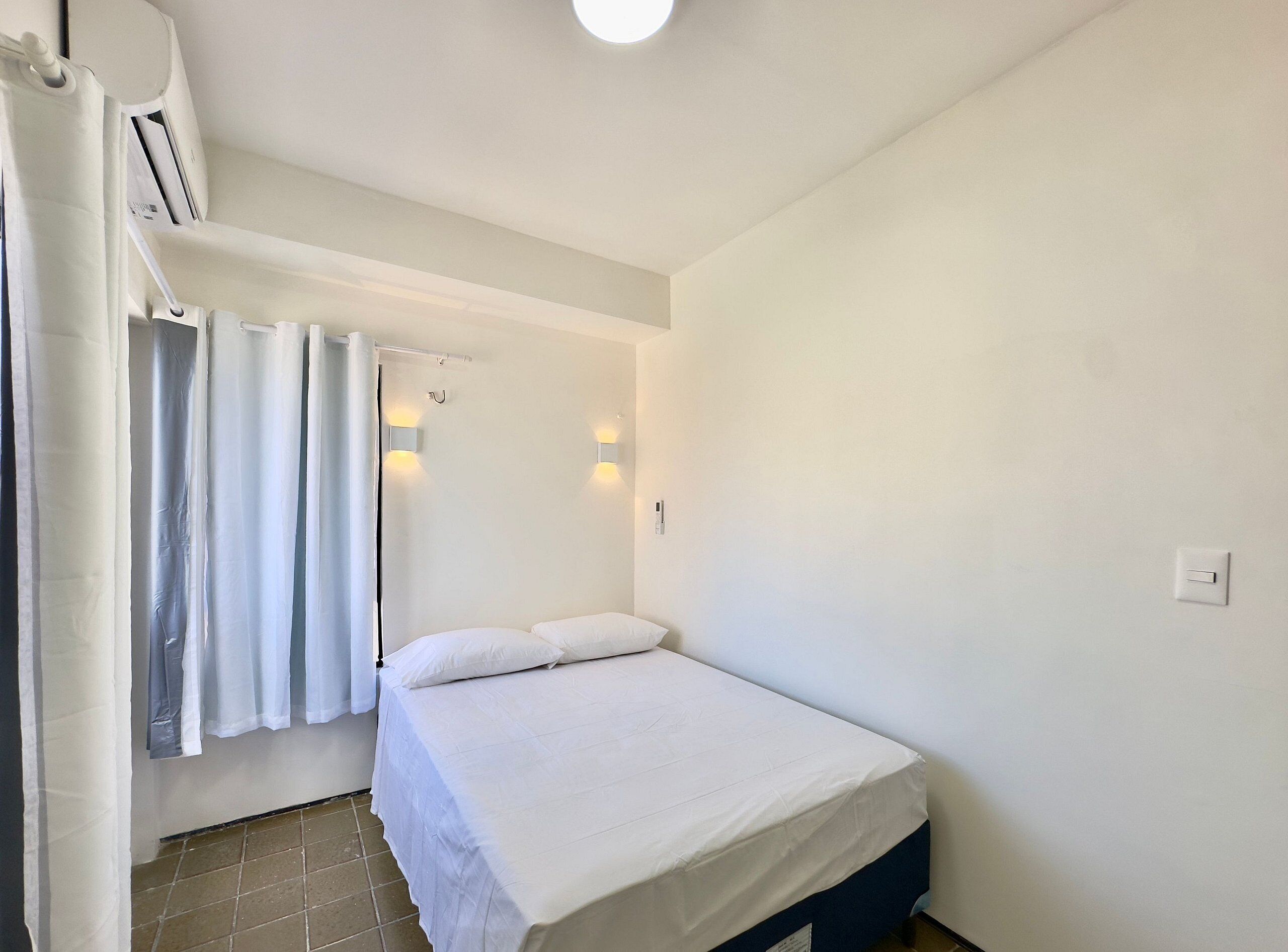 Una piccola camera con un letto singolo, un lenzuolo bianco, una tenda, una lampada a parete e una griglia di aerazione al soffitto.