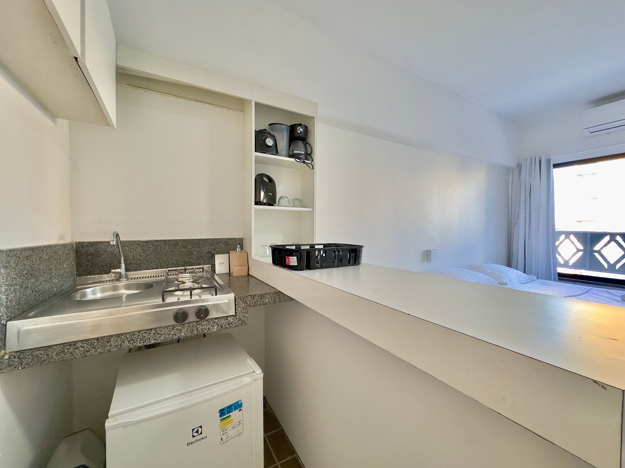 Una kitchenette con estufa a gas, pileta y heladera pequeña. Hay repisas sobre la mesada con diversos objetos.