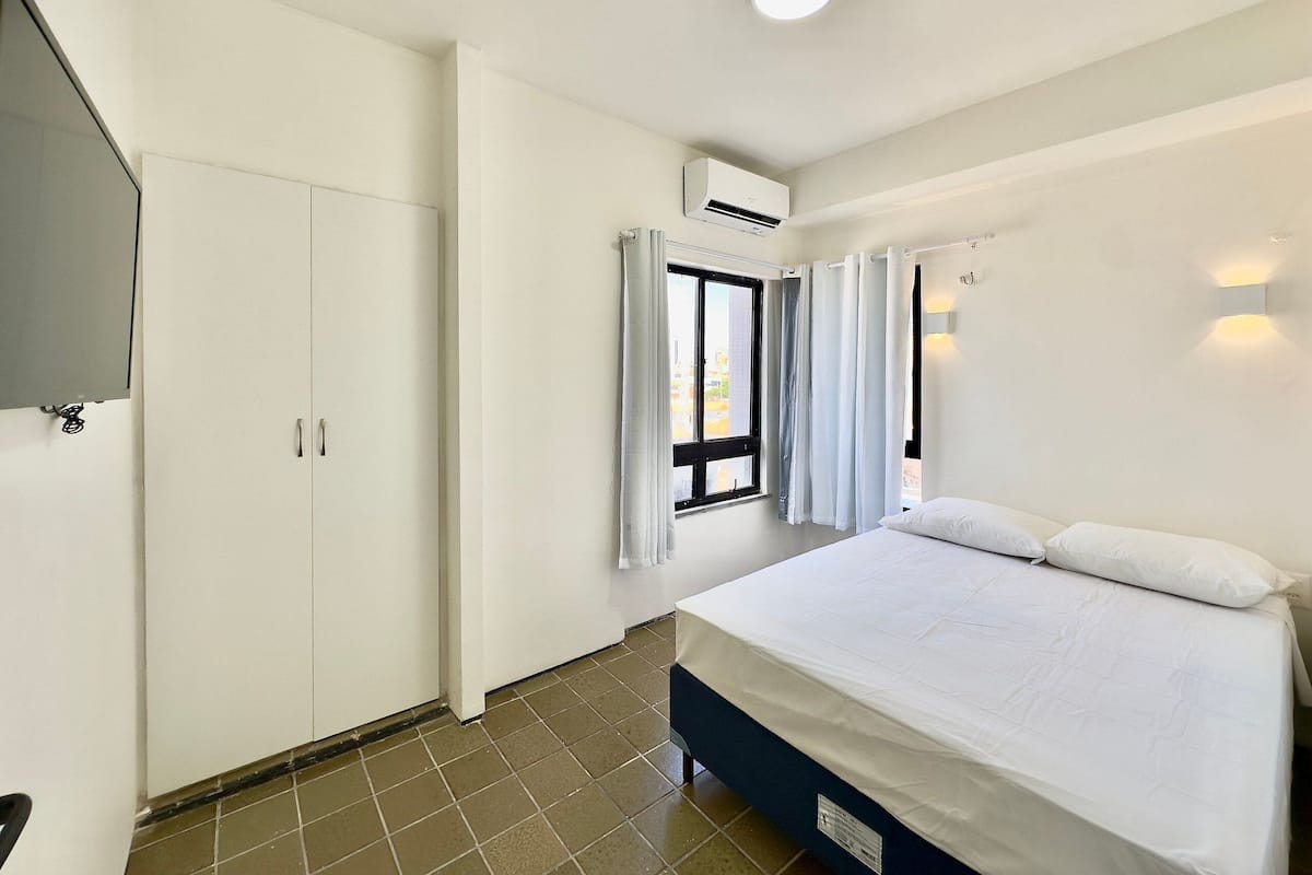 A bedroom with a bed, a wardrobe, an air conditioning unit, a window with curtains, and a wall-mounted light fixture.