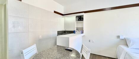 A compact room with a round table, two chairs, a bed, a kitchenette, and a small shelf.