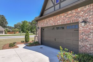 A residential garage with a brick facade, a garage door, and a small garden area with shrubs and a flower bush. - Peaceful AU Club Townhome / 3 Bed 3.5 Bath (Auburn)