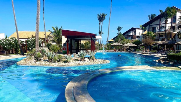 A swimming pool with a curved edge, palm trees, and a building with a red roof. - Taiba Beach Resort por Be My Guest! (São Gonçalo do Amarante)