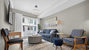 Ein modernes Wohnzimmer mit Sofa, Hocker, Sesseln, einem Fernseher und einem großen Fenster mit Vorhängen.