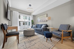 Ein modernes Wohnzimmer mit Sofa, Hocker, Sesseln, einem Fernseher und einem großen Fenster mit Vorhängen.