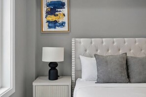 Un dormitorio con cabecera tapizada blanca, una mesita de noche con lámpara y un cuadro abstracto enmarcado en la pared.