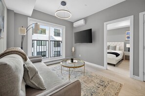 Ein modernes Wohnzimmer mit einer Couch, einem Couchtisch und einem Fernseher. Durch das Fenster ist ein Balkon zu sehen.