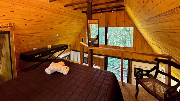 Une pièce de cabane en bois comprenant un lit, un banc, un bureau et une fenêtre donnant sur la forêt.