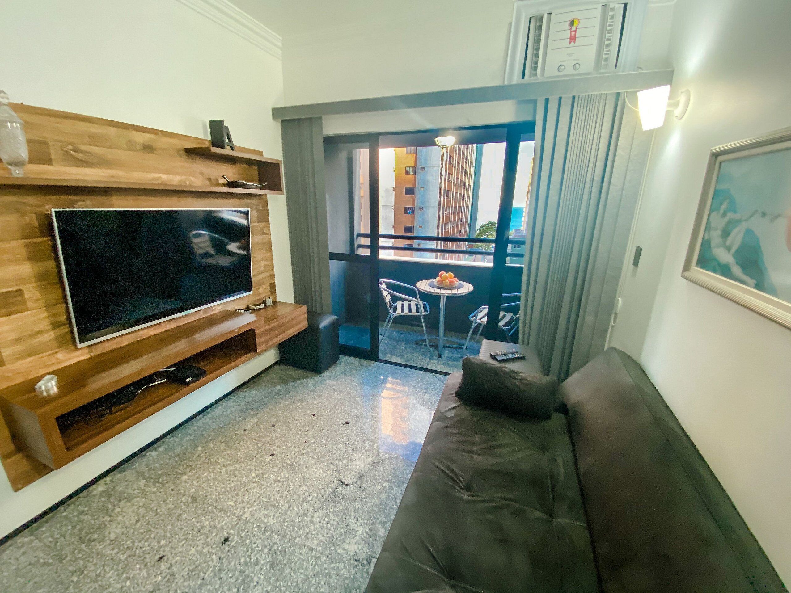 A living room with a TV on a wooden stand, a sofa, a small table with fruit, and a balcony with chairs.