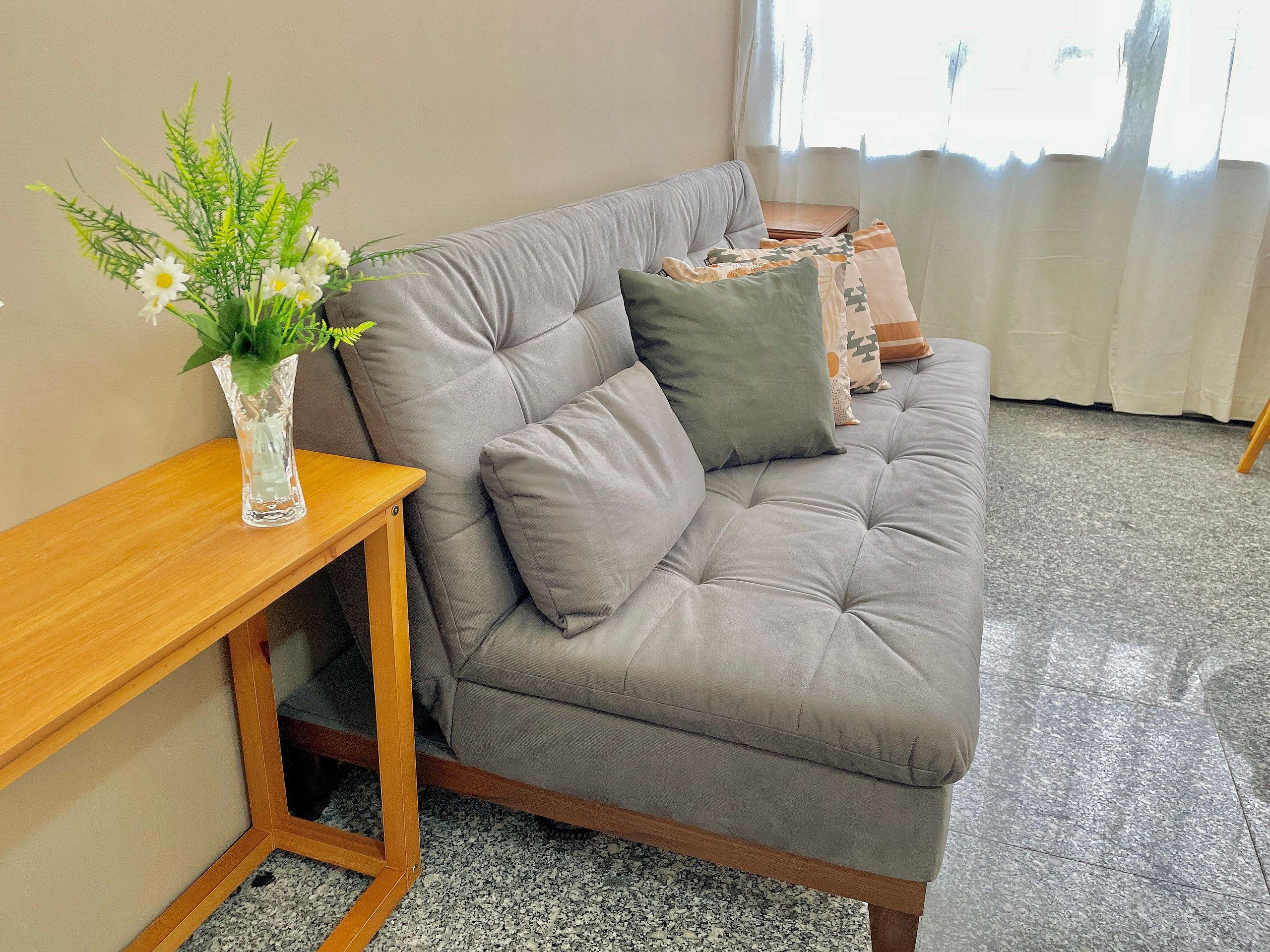A grey sofa with a cushion, a wooden side table, and a clear vase with flowers.