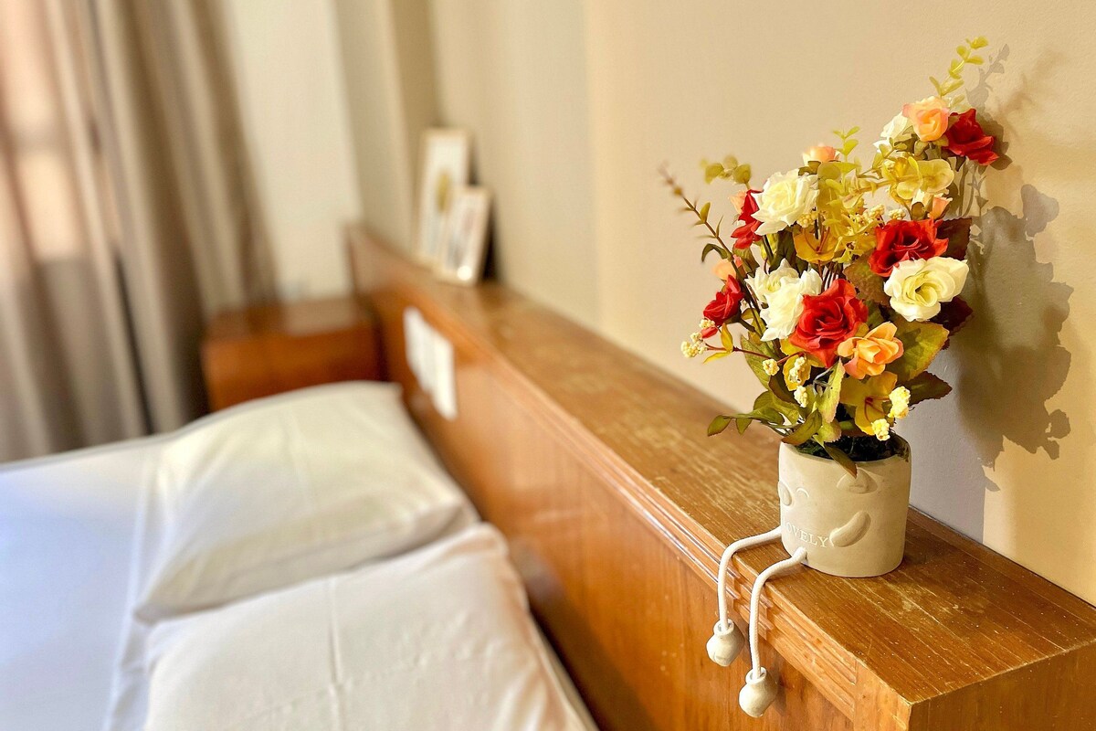 A hotel room with a bed, a wooden headboard, a wall-mounted vase with flowers, and a picture frame.