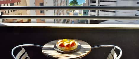 A balcony with a table and chairs, a bowl of fruit, and a view of the ocean and buildings.