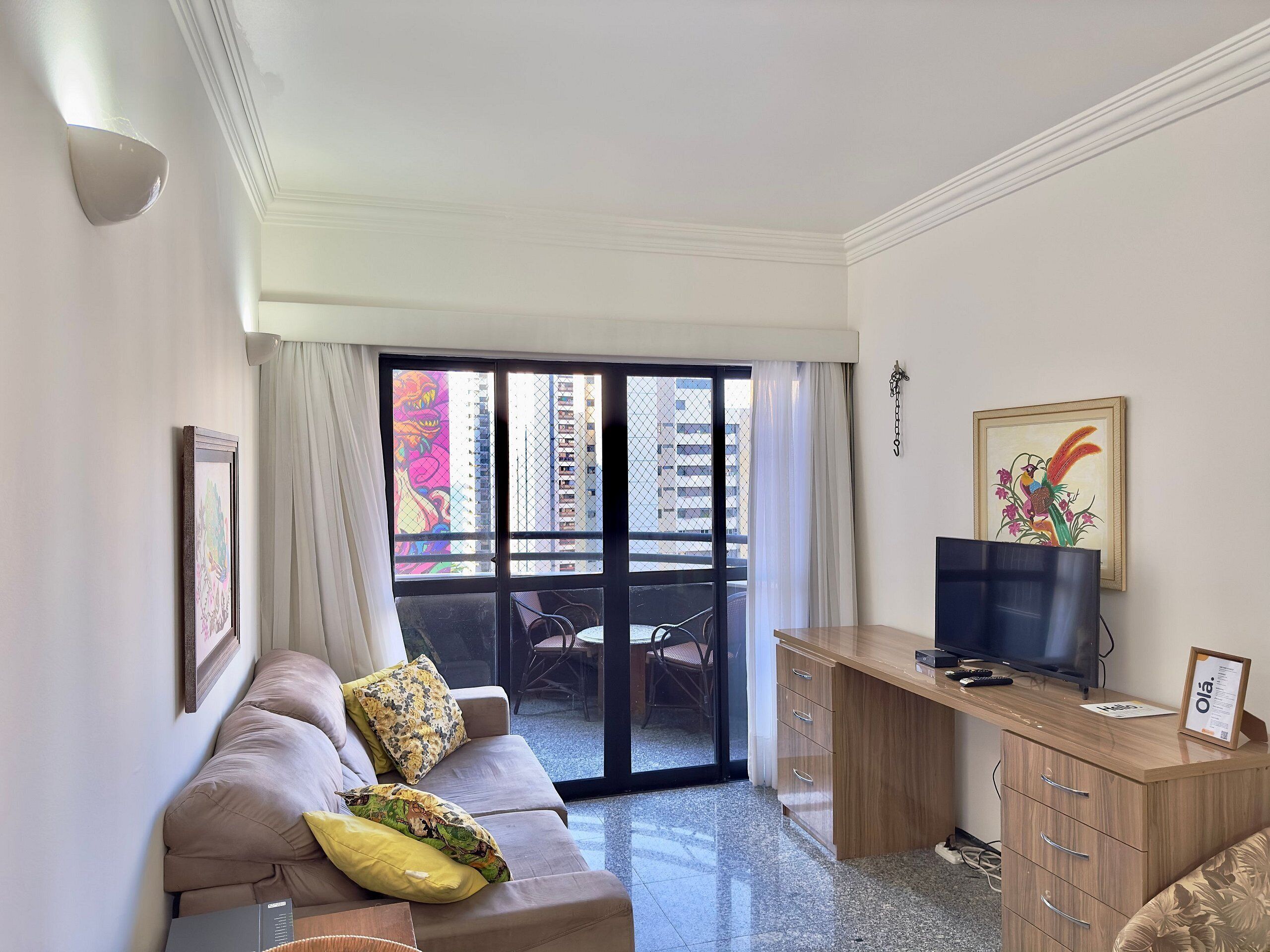 A living room with a sofa, a TV, a desk, and a balcony with a view of buildings.