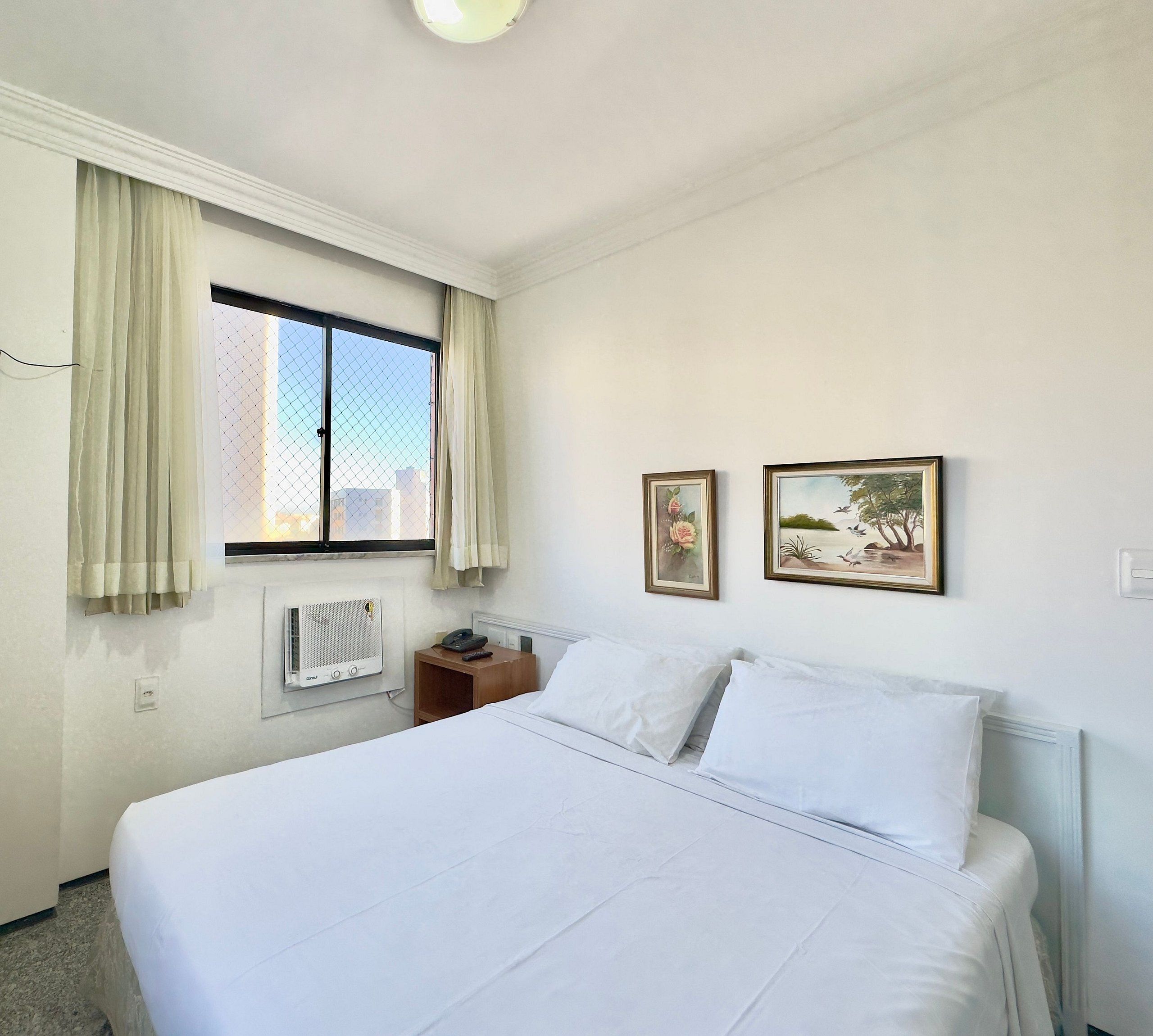 Ein Hotelzimmer mit einem Bett, einem Nachttisch, einem Fenster mit Vorhängen, einer Klimaanlage und zwei gerahmten Bildern an der Wand.