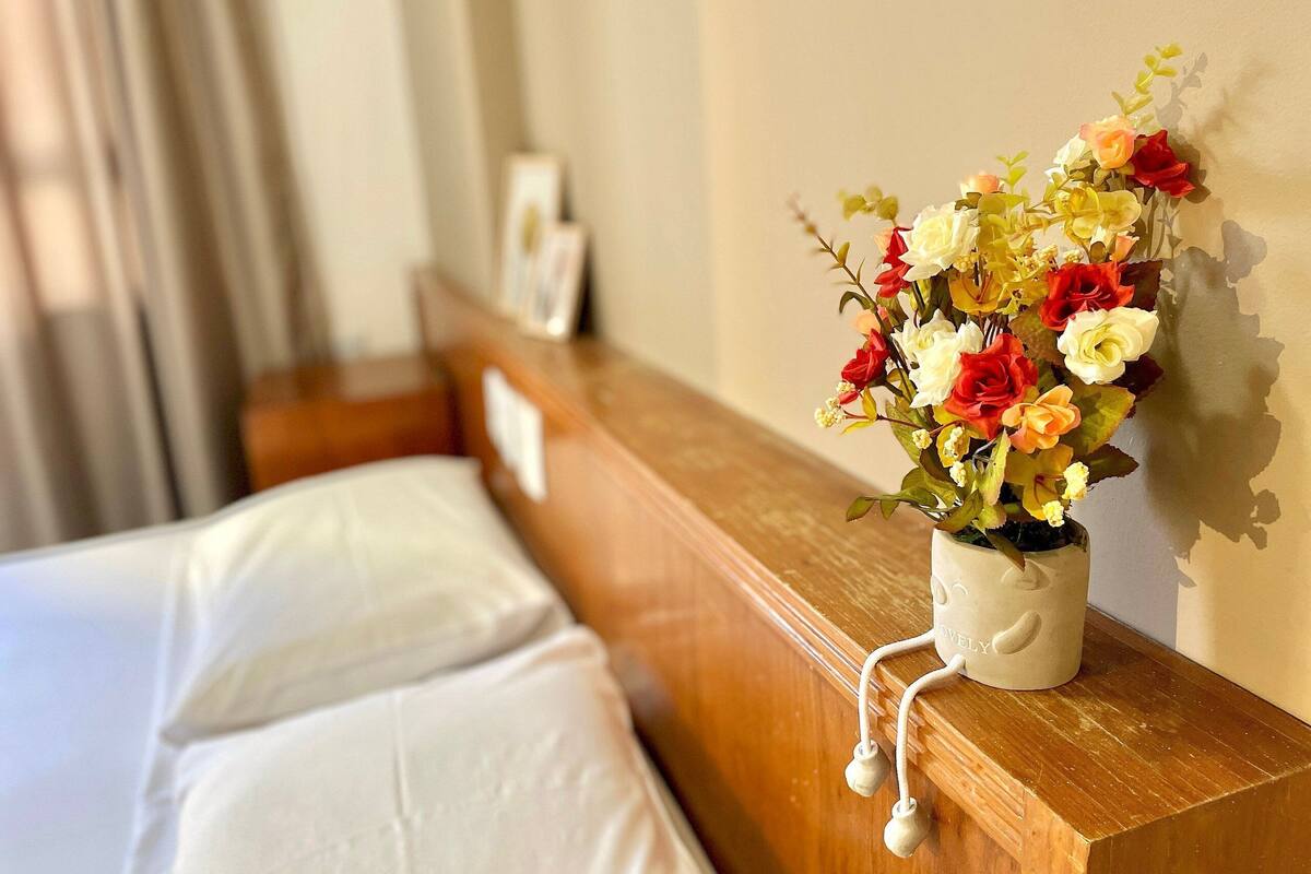 A hotel room with a bed, a wooden headboard, a wall-mounted vase with flowers, and a picture frame.