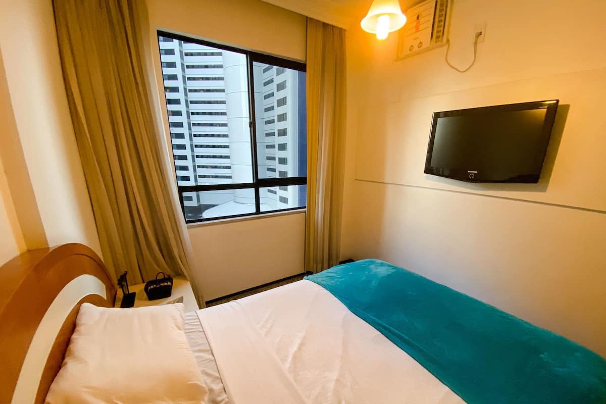 A hotel room with a bed, a TV mounted on the wall, a window with curtains, and a view of buildings outside.