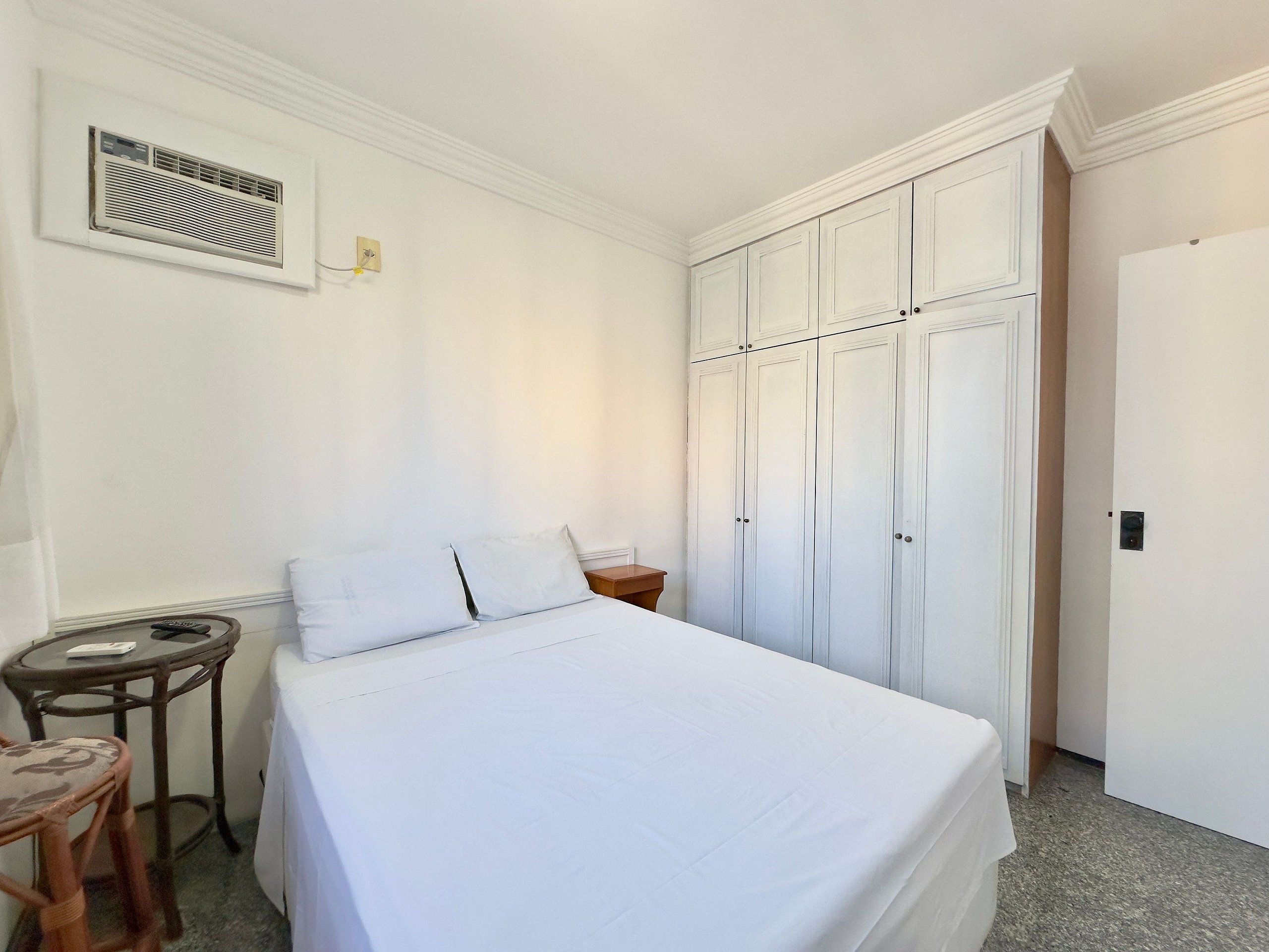 A bedroom with a bed, a small table, a chair, a wardrobe, and a wall-mounted air conditioning unit.